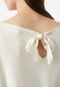 Kremowy dzianinowy top z wycięciem na plecach, zapinany na jedwabną kokardę. Dekolt lekko prążkowany, o gładkiej fakturze.