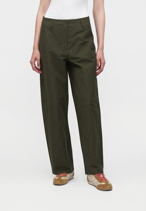 Mannequin portant un pantalon droit vert foncé et des baskets beiges avec des lacets rouges, debout devant un fond blanc uni.