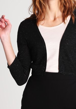 Femme portant un cardigan noir court par-dessus un top blanc et une jupe noire, avec des cheveux roux ondulés aux épaules, main levée près de l'épaule.