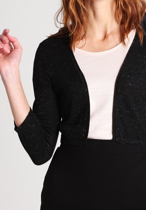 Femme portant un cardigan noir court par-dessus un top blanc et une jupe noire, avec des cheveux roux ondulés aux épaules, main levée près de l'épaule.