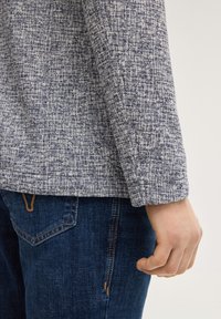 Persoon die een blauwe, getextureerde jas en donkerblauwe jeans draagt, met de rechterhand ontspannen langs het lichaam.