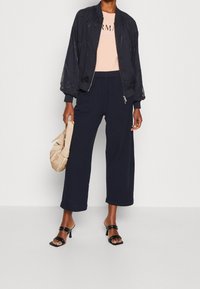 Černá bomber bunda s jemným vzorem, světle růžový top, tmavě modré široké kalhoty a černé sandály na vysokém podpatku s pásky. Bežová taška je součástí.