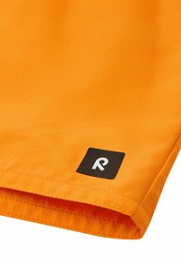 Pomarańczowy materiał o gładkiej fakturze, z zagiętym brzegiem oraz czarną kwadratową metką z białym logo w dolnym rogu.