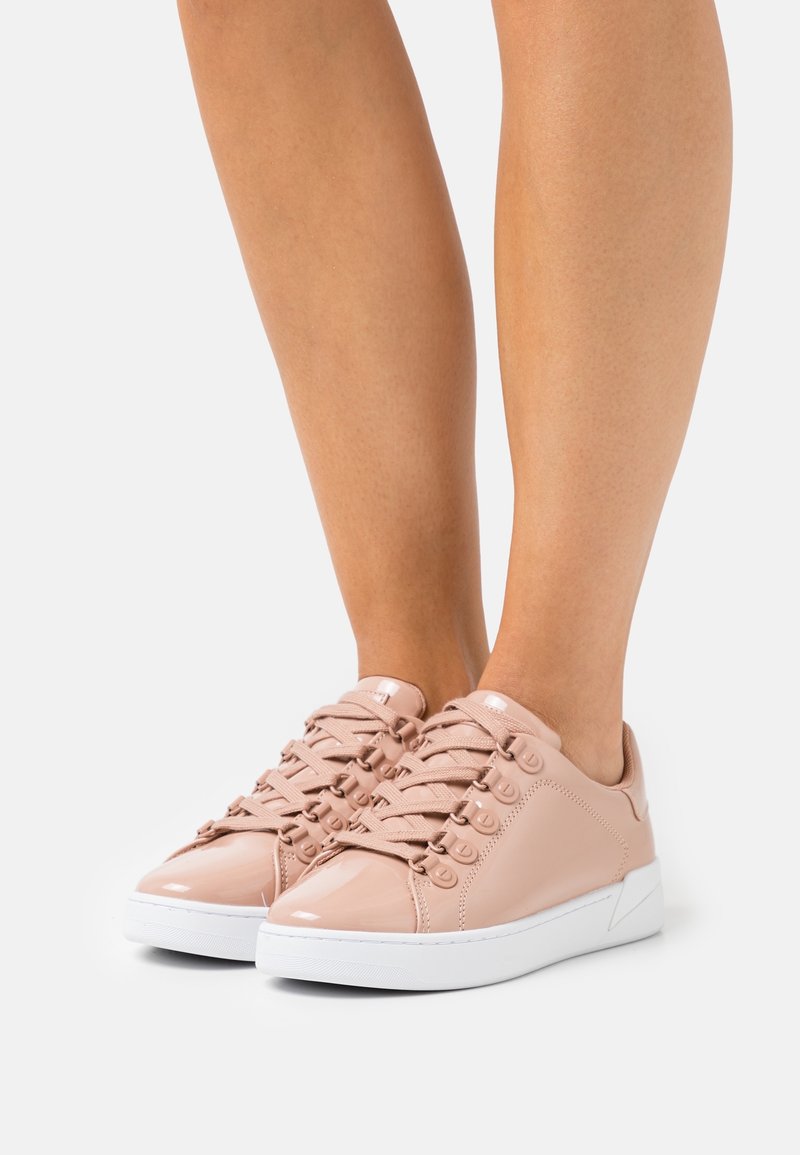 Jambes portant des baskets basses rose clair brillantes avec semelles blanches, lacets et bout rond sur fond blanc uni.