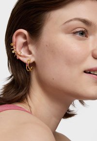 Orecchini color oro con gemme colorate, disponibili in diversi design tra cui anelli e perni, indossati all'orecchio di un modello.