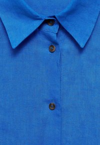Camicia in lino blu con colletto classico e tre bottoni marroni sul davanti, che evidenzia una texture liscia e un design semplice.
