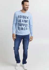 Camisa de manga larga azul claro con texto gráfico en azul oscuro, combinada con jeans azul oscuro y zapatillas blancas. Estilo casual.