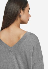Grau V-Ausschnitt Pullover mit lockerer Passform, weicher Textur und sichtbaren Nähten entlang des Halsausschnitts. Die Haare sind glatt getragen, sodass die Ohrringe sichtbar sind.