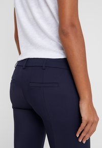 Personne portant un pantalon bleu marine ajusté et un haut blanc, montrant le dos et le côté avec une main reposant près de la cuisse sur un fond uni.