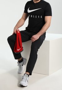 Hombre sentado en un bloque blanco vestido con una camiseta negra de Nike "ATHLETE", pantalones negros, zapatillas grises de Nike, sosteniendo una toalla roja de Nike con la mano izquierda.