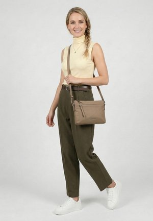 Young woman with braided hair wears sleeveless beige top, olive pants, white sneakers, and carries a beige crossbody bag, smiling.