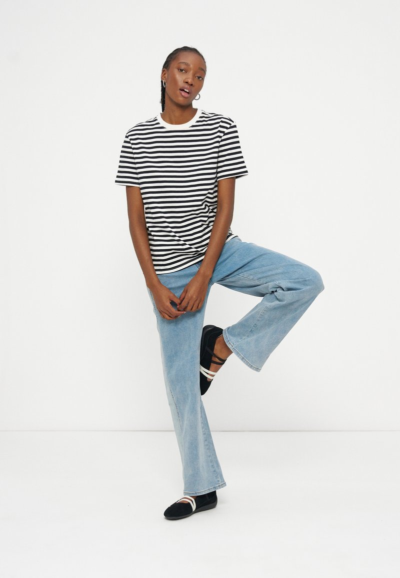 Jeune personne portant un t-shirt rayé noir et blanc, un jean bleu clair, des ballerines noires, debout sur une jambe sur un fond blanc uni.