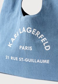 Sacolinha de denim com uma alça curva, tecido azul claro, e texto impresso em branco: "KARL LAGERFELD PARIS 21 RUE ST-GUILLAUME."