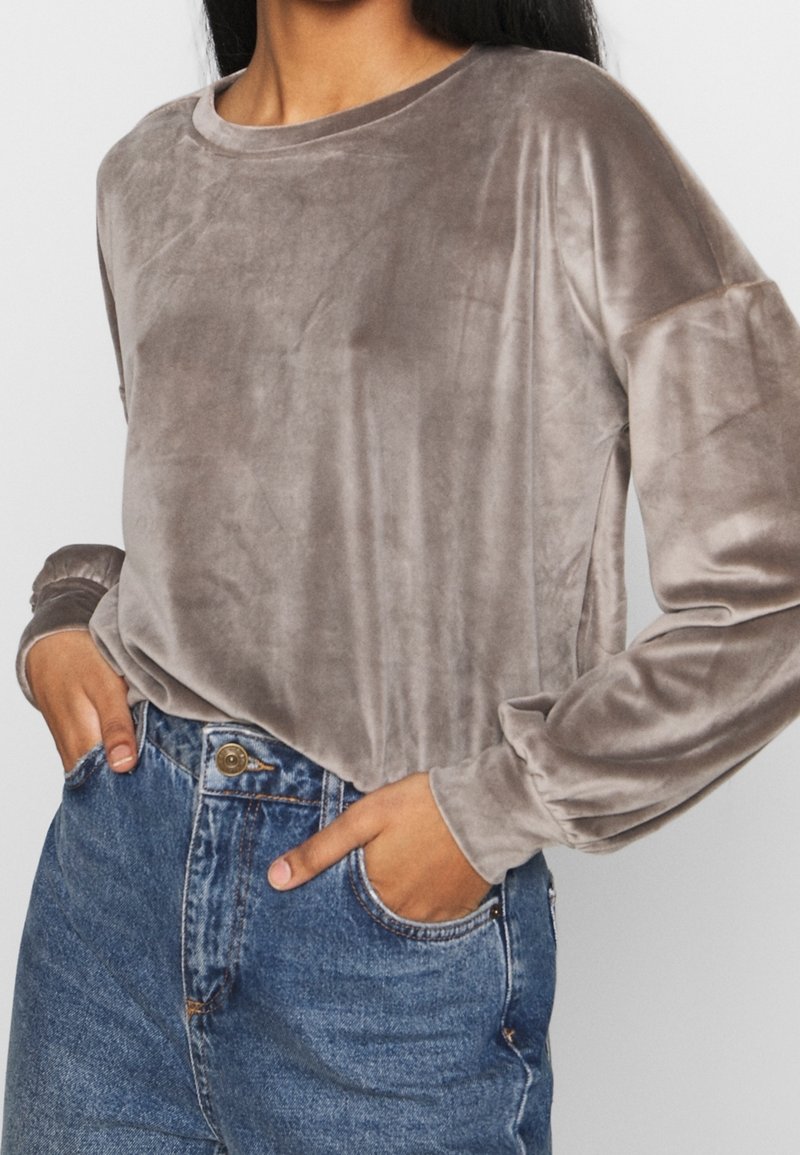 Personne portant un haut ample à manches longues en velours gris rentré dans un jean bleu taille haute, les mains dans les poches.