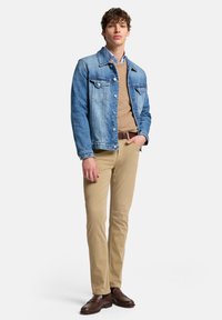 Młody mężczyzna ubrany w dżinsową kurtkę, jasnobrązowy sweter na niebieską koszulę, beżowe spodnie i brązowe mokasyny, stoi z jedną ręką w kieszeni.