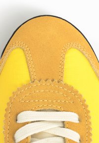 Gros plan sur l'avant jaune d'une chaussure de sport avec des empiècements en daim beige et des lacets blancs sur fond blanc.
