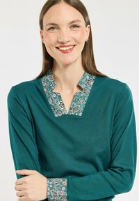 Haut à manches longues teal avec un col en V et un col et des poignets à motifs comportant des designs floraux dans des tons de vert, violet et rose.