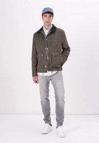 Olive green jacket with a collar, featuring multiple front pockets and button closure. Worn with light gray jeans and white sneakers.