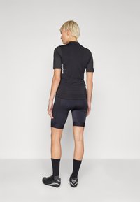 Schwarzes Radtrikot mit kurzen Ärmeln, hat ein enges Design, eine glatte Textur und ein Logo auf der linken Seite. Schwarze gepolsterte Radhosen.