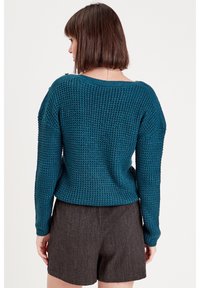Pull en tricot teal avec une coupe ample, un large col et un motif texturé. Associé à un short gris foncé. Doté de détails en bouton sur l'épaule.