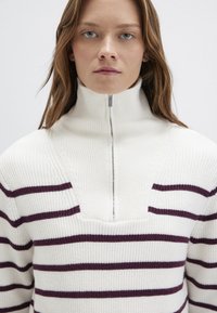 Weißer Strickpullover mit dunkelrot gestreiften horizontalen Linien, hohem Kragen und einem halben Reißverschluss auf der Vorderseite. Strukturiertes Rippmuster.
