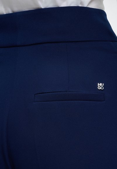 Gros plan sur un pantalon bleu marine avec une poche passepoilée à l’arrière et un petit logo métallique LIU JO au-dessus de la poche.