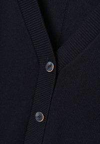Gros plan sur un cardigan noir en maille avec un col en V côtelé et deux boutons noirs bordés de beige attachés.