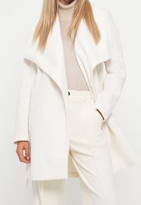 Vit ullblandad kappa med vattenfallskrage, framfickor och skräddarsydd passform, i kombination med en beige polotröja och matchande byxor.