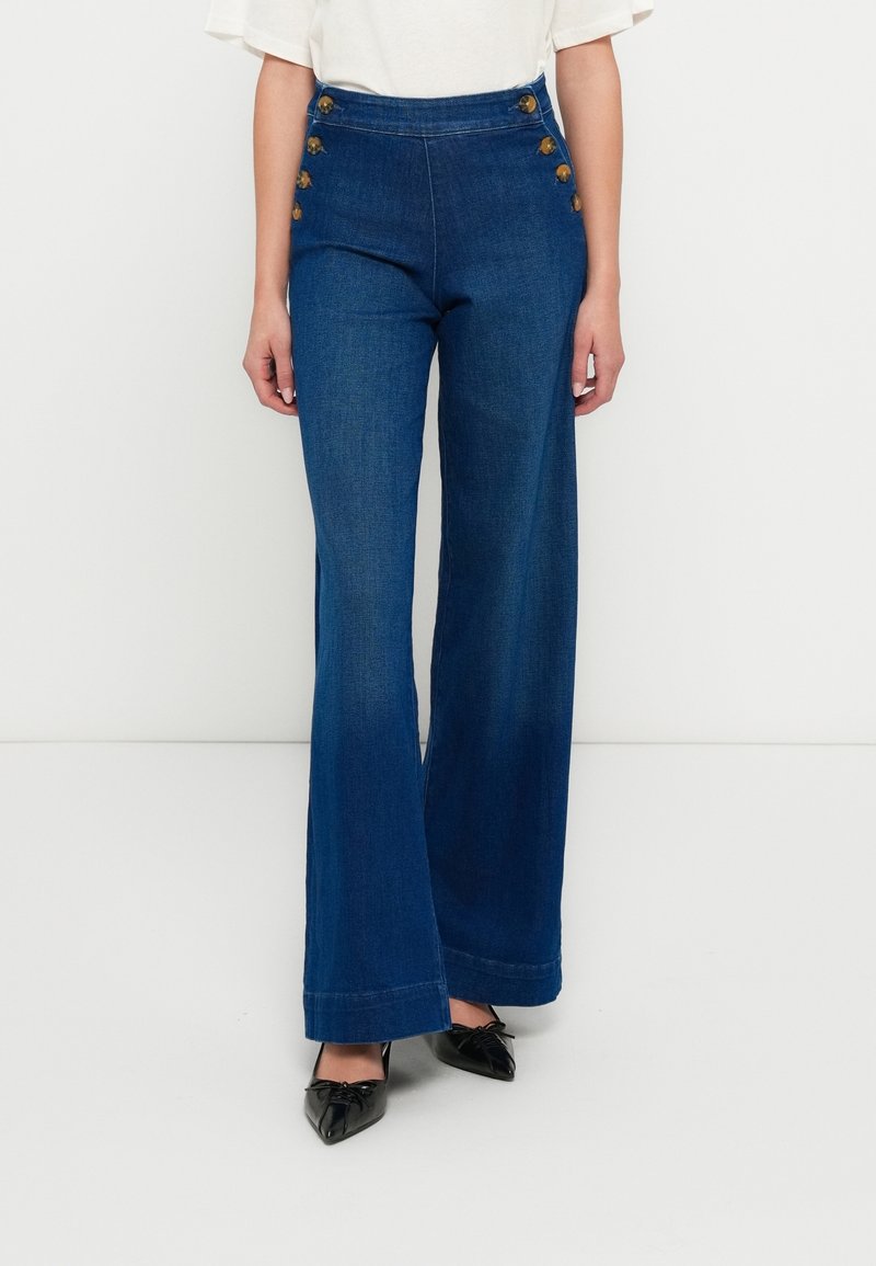 Femme portant un jean bleu taille haute à jambes larges avec des boutons le long de la taille, associé à des chaussures noires à bout pointu et un haut blanc.