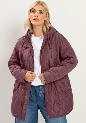 Femme portant une veste matelassée à capuche bordeaux sur une chemise blanche et un jean bleu sur un fond neutre.