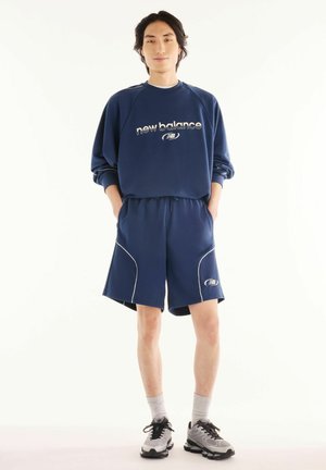 Una giovane persona è in piedi con le mani nelle tasche, indossando una felpa blu navy New Balance e pantaloncini coordinati con sneakers e calzini grigi.