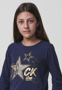 Tamsiai mėlyna marškiniai ilgomis rankovėmis su metalinėmis aukso žvaigždėmis ir tekstu "CK žvaigždė". Pagaminta iš minkštos medvilnės medžiagos ir suapvalintu kaklu.