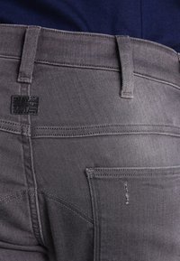 Primer plano de unos jeans de mezclilla gris que se llevan con una camisa azul marino, mostrando el bolsillo trasero, los lazos del cinturón y una etiqueta de marca negra en la cinturilla.