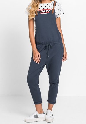 Salopette en coton bleu marine avec bretelles marron réglables, taille à cordon de serrage et poches latérales. Associée à un t-shirt blanc à pois et des baskets.