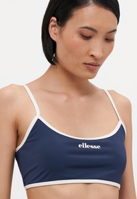 Mørkeblå bh med tynne hvite stropper, elastisk materiale, og en hvit "ellesse" logo sentrert foran. Glatt tekstur.