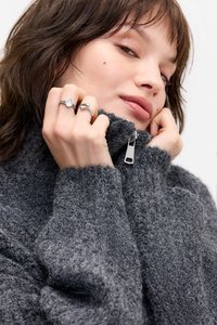 Pulôver cinza texturizado com gola alta e fecho de zip parcial. O modelo exibe anéis prateados, realçando as mãos e acessórios.