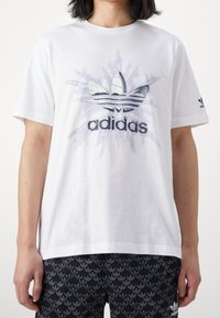 Bílý bavlněný tričko s centrálním grafickým motivem loga Adidas s efektem rozbití. Krátké rukávy, kulatý výstřih.