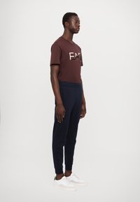 Barna, rövid ujjú póló grafikus szöveggel, sötétkék melegítőnadrággal és fehér sportcipővel párosítva. A nadrág rugalmas külső szegéllyel és derékrésszel rendelkezik.