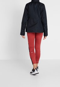 Zwarte waterdichte jas met lange mouwen, gecombineerd met strakke rode leggings met zwarte zijpanelen, en zwarte sportschoenen.