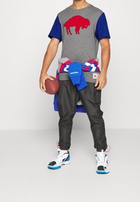 Grå t-shirt med rød bøffelgrafik, blå ærmer; sorte joggers; lagde blå og røde sweatshirts om livet; brun fodbold; hvide sneakers.