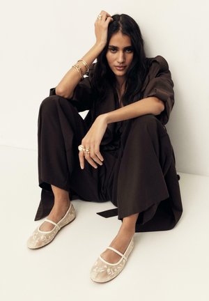 Vrouw met lang donker haar, gekleed in losse donkere kleding en beige Mary Jane-schoenen met borduursel, zittend tegen een effen witte muur.