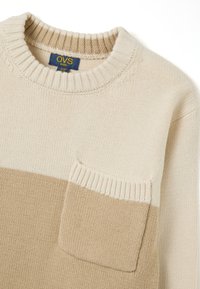 Suéter de punto beige con una sección superior en crema. Presenta un cuello y puños acanalados, y un bolsillo de parche texturizado en el lado izquierdo.