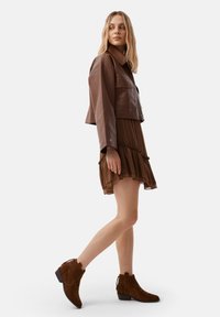 Giacca cropped in pelle marrone sopra un abito marrone plissettato, abbinata a stivali in suede marrone al polpaccio, con tacco a blocco.