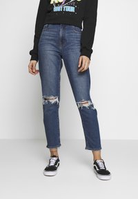 Högmidjade slitna blå jeans med hål vid knäna, avslutade med en rå fåll, kombinerade med svarta sneakers och en grafisk svart sweatshirt.