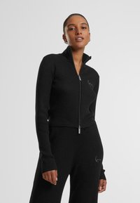 Pull noir côtelé zippé avec col montant, design ajusté, manches longues et fermeture éclair argentée. Présente des détails brodés discrets.