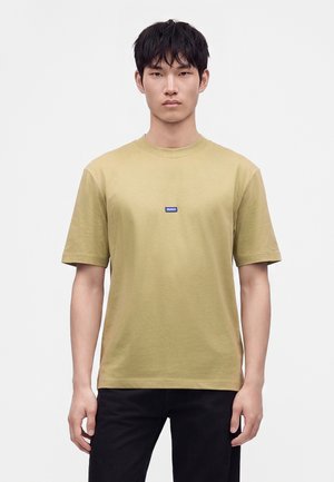 Jeune homme aux cheveux noirs courts portant un t-shirt uni marron clair avec un petit logo bleu Hugo sur la poitrine et un pantalon noir.