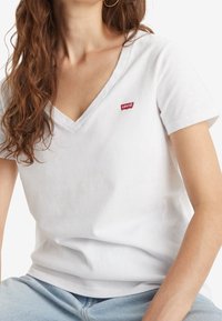 Femme aux longs cheveux bruns portant un t-shirt blanc Levi's à col en V et un jean bleu clair, assise devant un fond blanc uni.