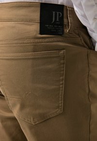Bruine katoenen broek met een gestructureerd oppervlak, zichtbare stiksels en een zwart etiket op de tailleband. Heeft een achterzak met een opvallend ontwerp.