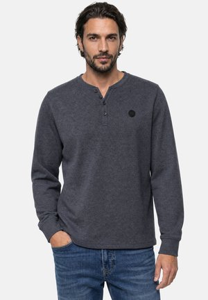 Homme aux cheveux foncés et barbe, portant une chemise henley gris foncé à manches longues et un jean bleu, une main dans la poche.