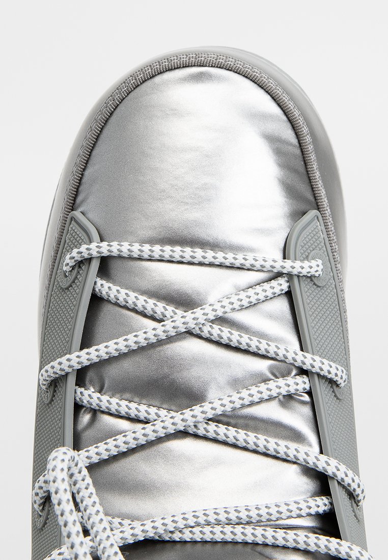 Silberner Sneaker mit weiß-grauen, gemusterten Schnürsenkeln, die über ein gepolstertes, glänzendes Obermaterial und graue Seitenstützen gekreuzt sind.